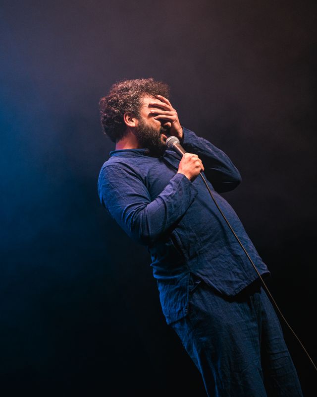 Nish stands on stage with a microphone with his face in his hands.