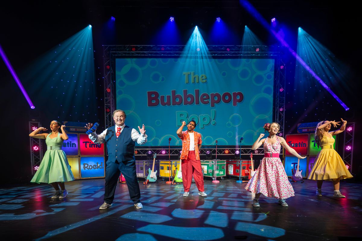 Justin holds a tambourine with the ensemble dancing around him.