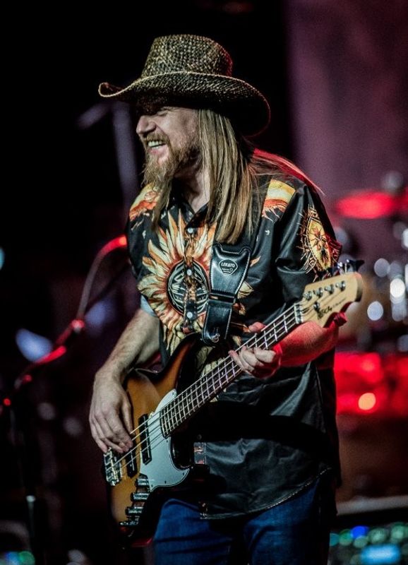 A person playing a bass guitar on stage, wearing a cowboy hat and a patterned shirt. The background features drums and stage lights creating a vibrant atmosphere.