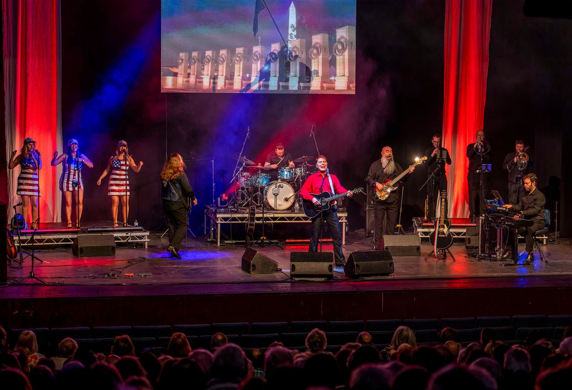 The band play on stage backed by three singers who are wearing dresses with the USA flag on.
