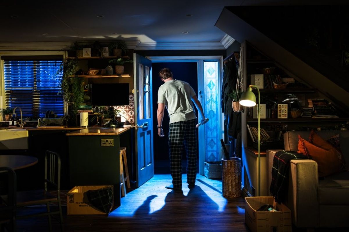 A man is looking out of the kitchen door into the outside. He is holding a bat.