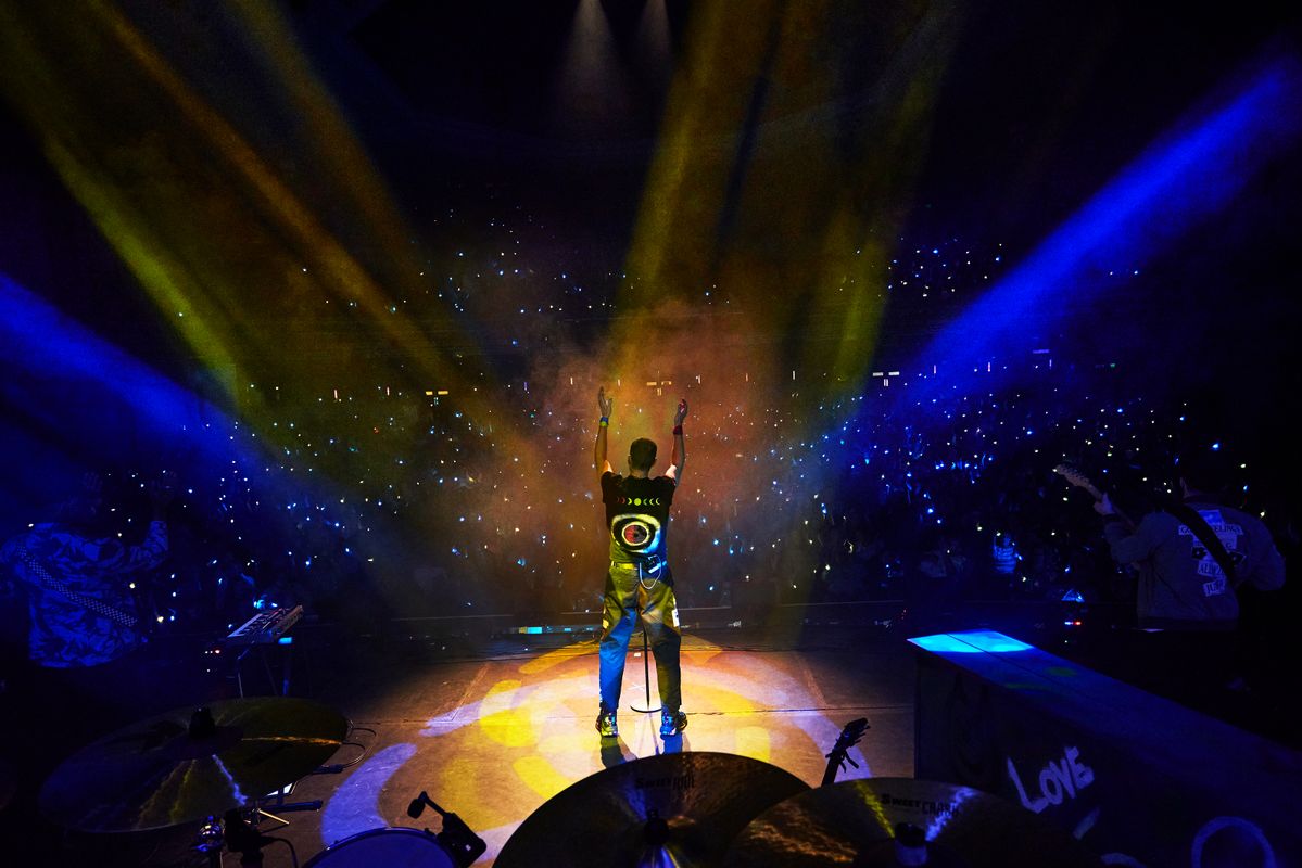 A performer stands on stage, arms raised, facing a cheering crowd. Vibrant blue and yellow lights illuminate the scene, creating an energetic and celebratory atmosphere.