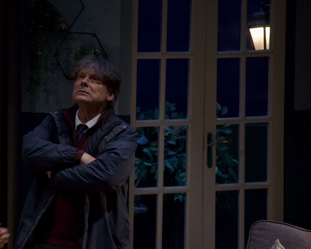 A person is stood in a dimly lit living room setting with their coat on 