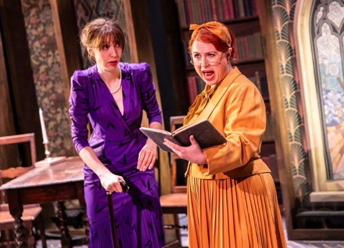 Two women in period costumes perform on stage. One, in a purple dress, holds a cane and looks concerned. The other, in orange, reads dramatically from a book.