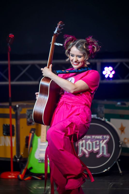 A cast member dressed head to toe in pink plays an acoustic guitar.