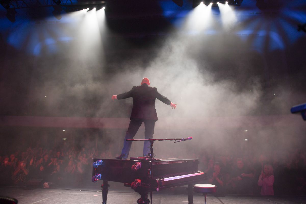 A person stands on a grand piano, surrounded by smoke, looking out into the audience.