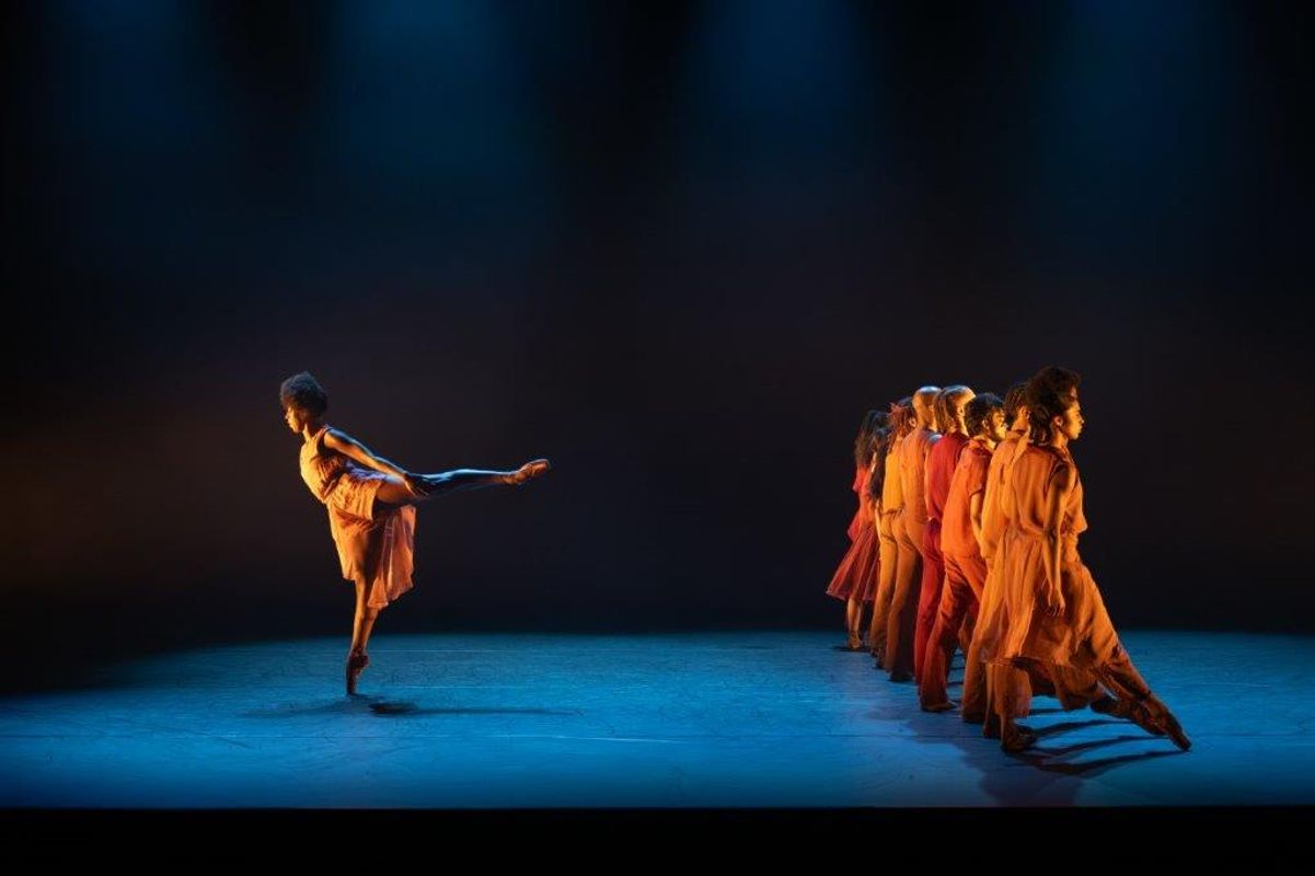 Group of dancers in orange and brown clothing with a soft spot light on them. One has their leg up behind them and the others are in a line facing away from them