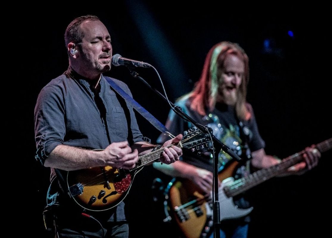 Two musicians perform on stage. The person in the foreground plays a mandolin and sings into a microphone. The background musician plays an electric bass guitar. The lighting is focused on the performers against a dark backdrop.