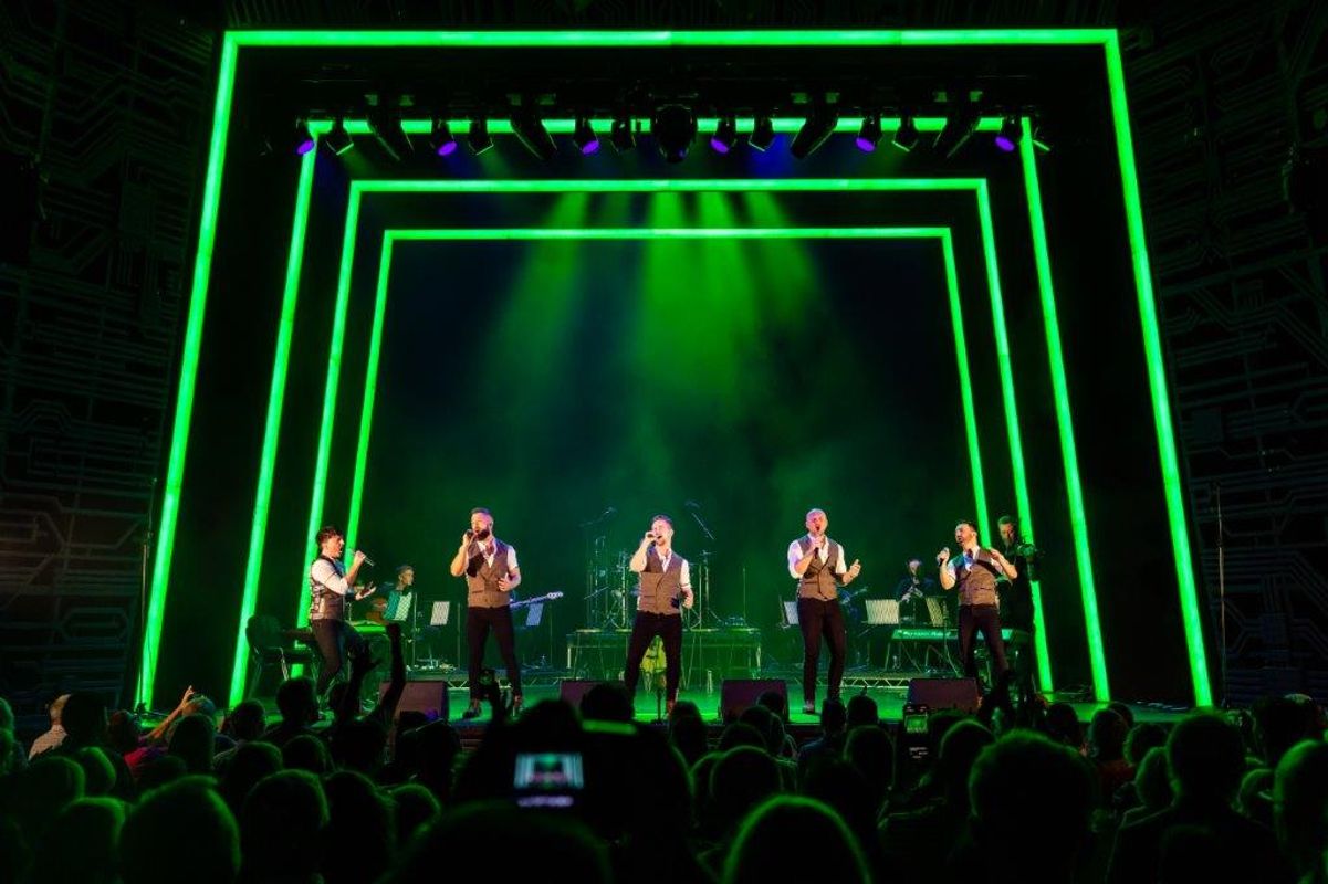 A lively band performing on stage under vibrant green lights with glowing frames. The crowd is engaged, capturing the energetic concert atmosphere.