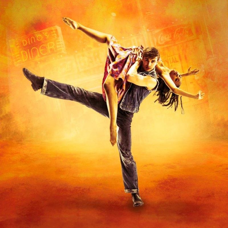 A dancer in denim holds a partner mid-air against an orange-yellow backdrop. The dynamic pose and vibrant background convey energy and passion.