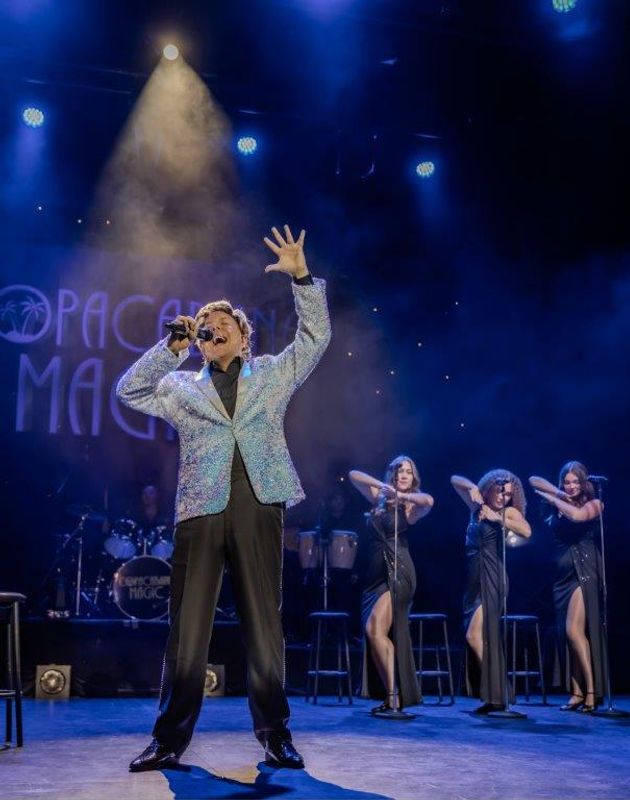Martyn James in a sparkling blazer energetically sings on stage. Three women in elegant black dresses dance behind him. Blue lights and smoke create a lively ambiance.