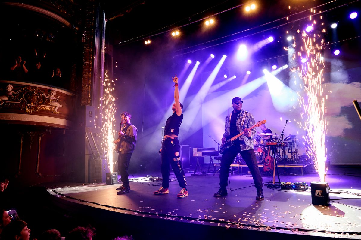 A vibrant band performs on stage, surrounded by colorful lights and fireworks. The lead singer raises a finger triumphantly, creating an energetic atmosphere.