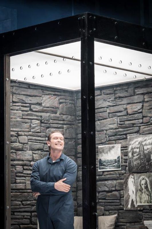 A man wearing navy overalls is stood in a glass cell. He has his arms folded and is smiling