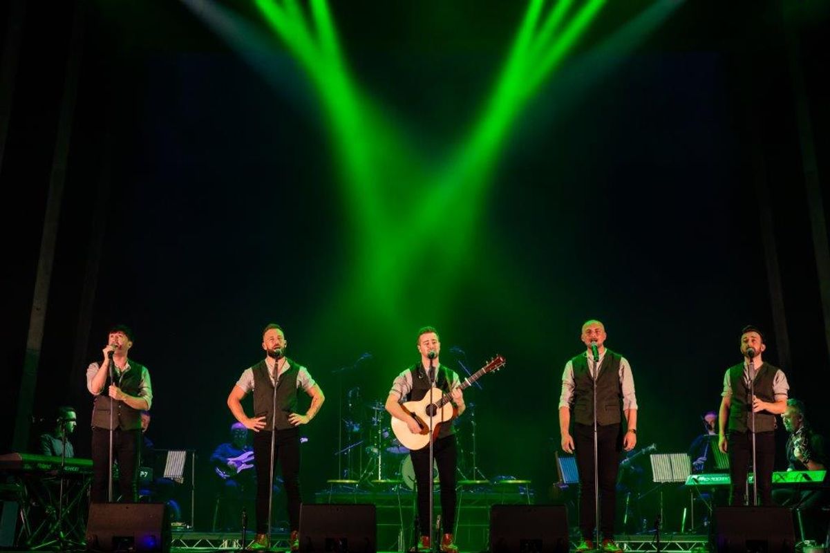 Five men (Shamrocks) perform on stage, each holding a microphone. One plays a guitar. They are illuminated by vibrant green stage lights, creating an energetic atmosphere.
