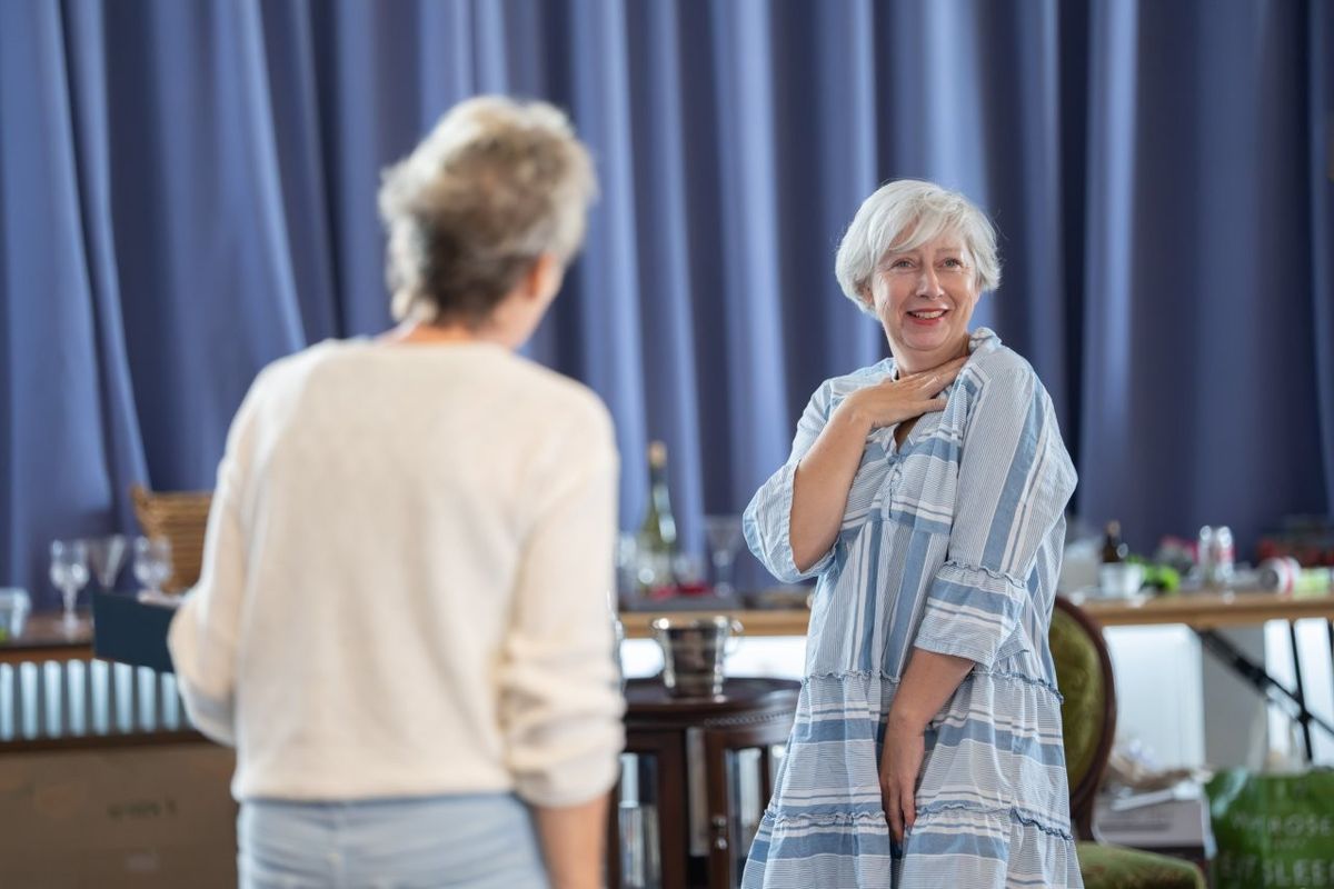 Two women are stood, one is facing the other, the other woman looks on smiling with her hand on her chest