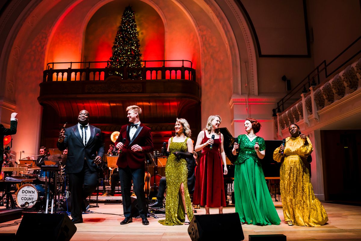Six people stand on stage singing with a Christmas Tree in the background.