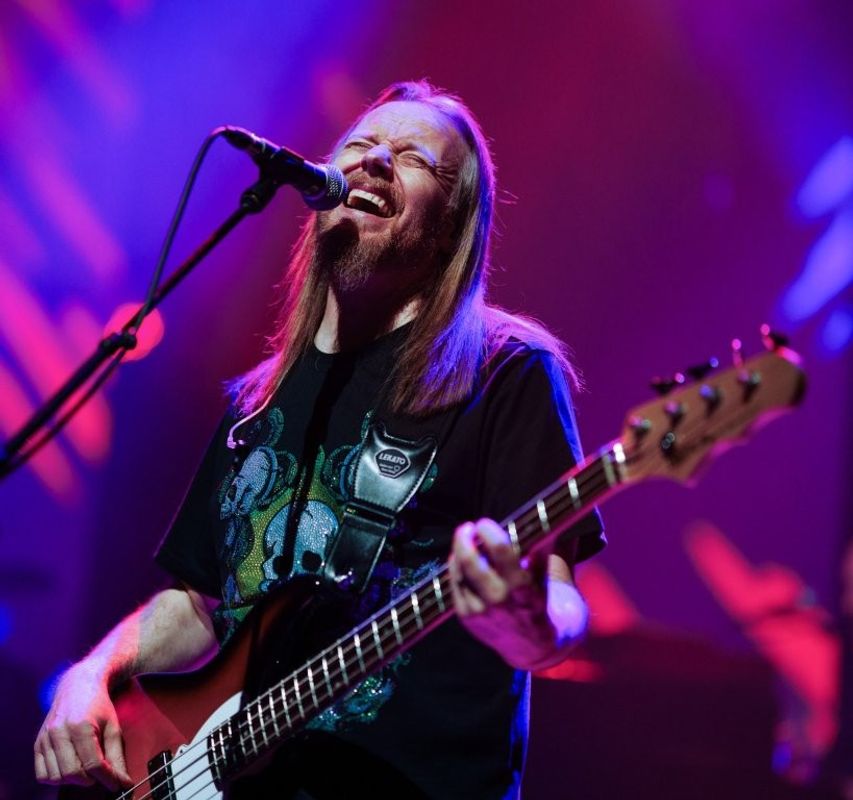Musician passionately playing a bass guitar and singing into a microphone on a vibrant stage with colorful lighting.