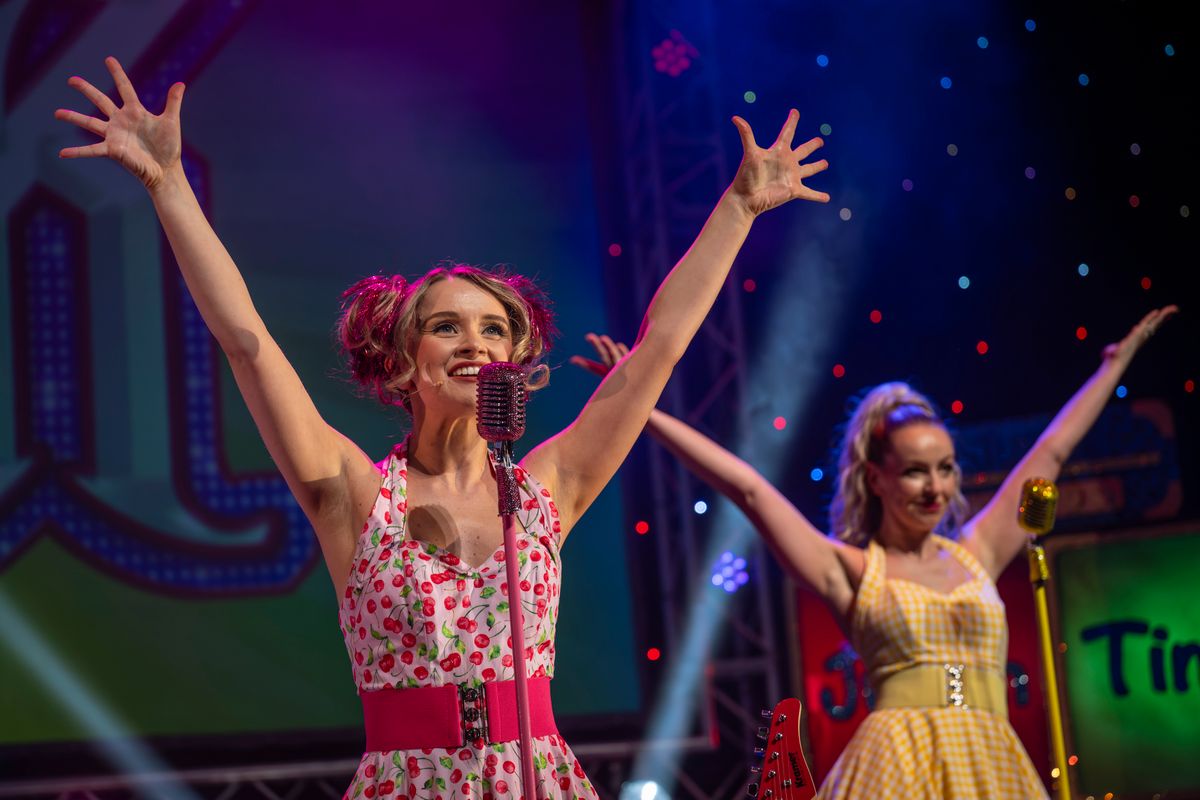 Two singers wearing 1950's inspired dresses stand with their arms up in the air.