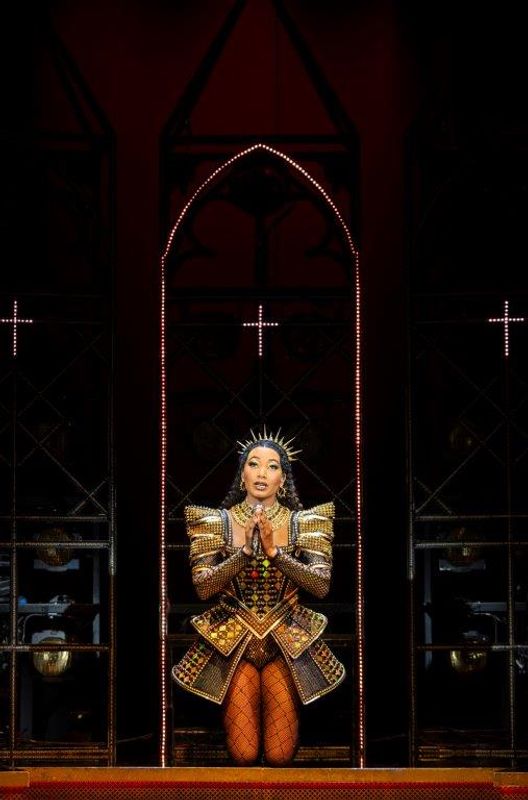 A person wearing a gold jewelled outfit and crown headband stands in front of a cross.