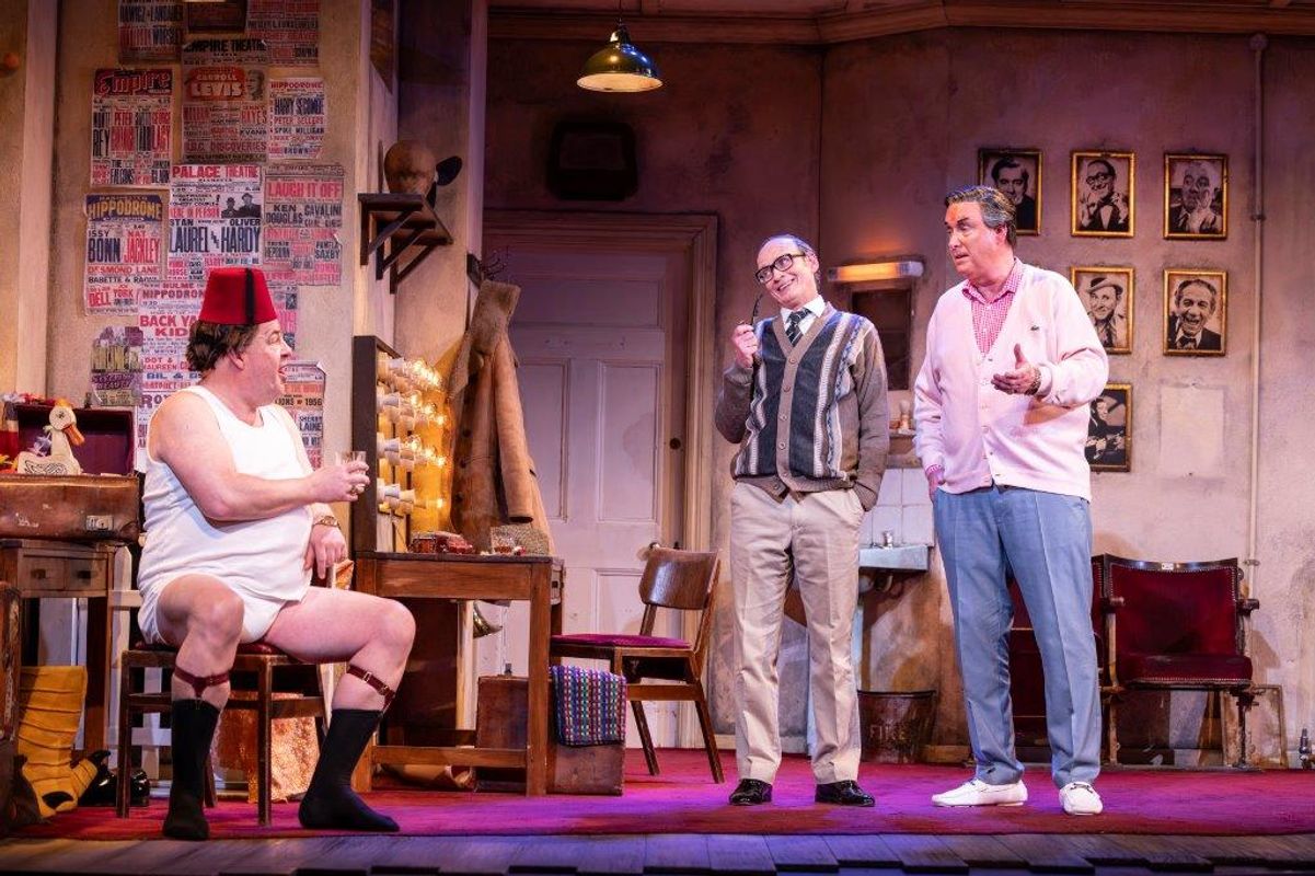 Three actors perform in a lively theater scene. One sits wearing a fez, another stands mid-laugh with glasses, and the third smiles in pastel clothing. The room is adorned with posters and framed photos. The setting is warmly lit, creating a comedic and vibrant atmosphere.