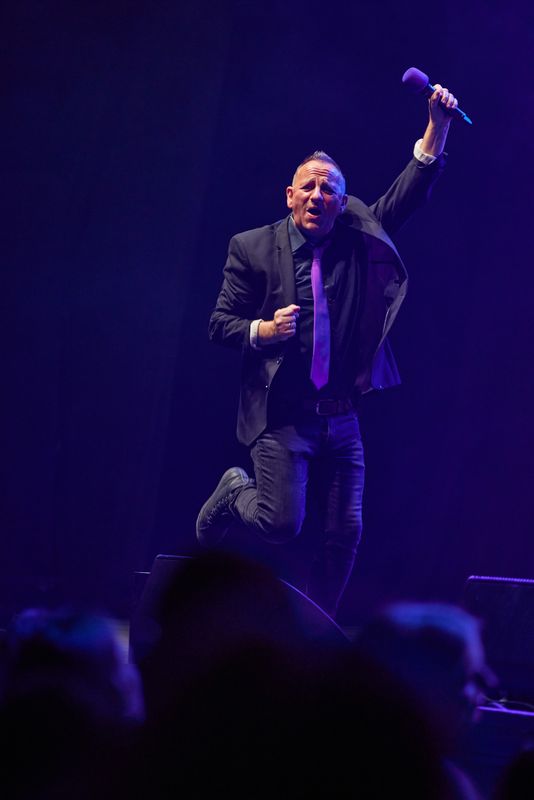 A person wearing a black suit and purple tie raises their left arm in the air.