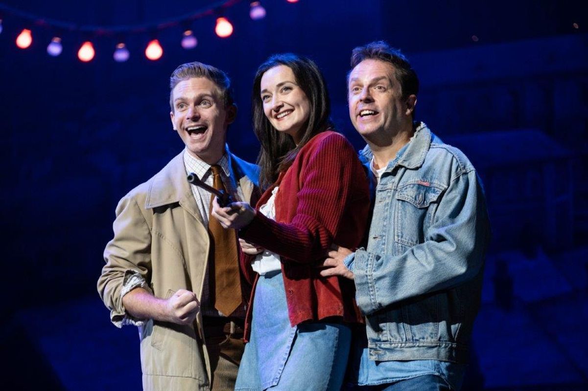 Three people are stood together, there is a woman in the middle and one man either side of her. They are all smiling