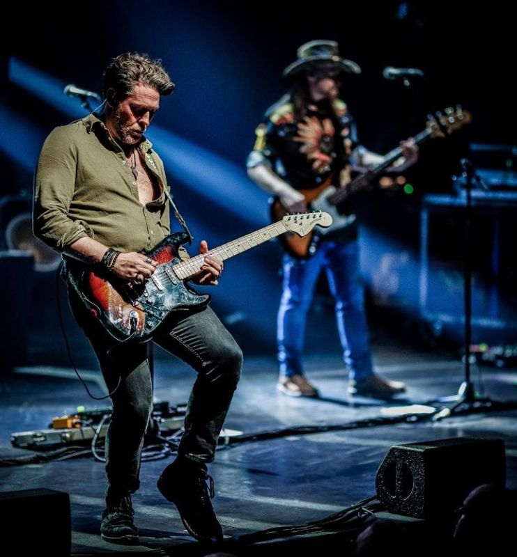 Two musicians performing on stage. The one in the foreground is playing an electric guitar, while the one in the background is playing a bass guitar. Bright stage lights create a dramatic atmosphere.