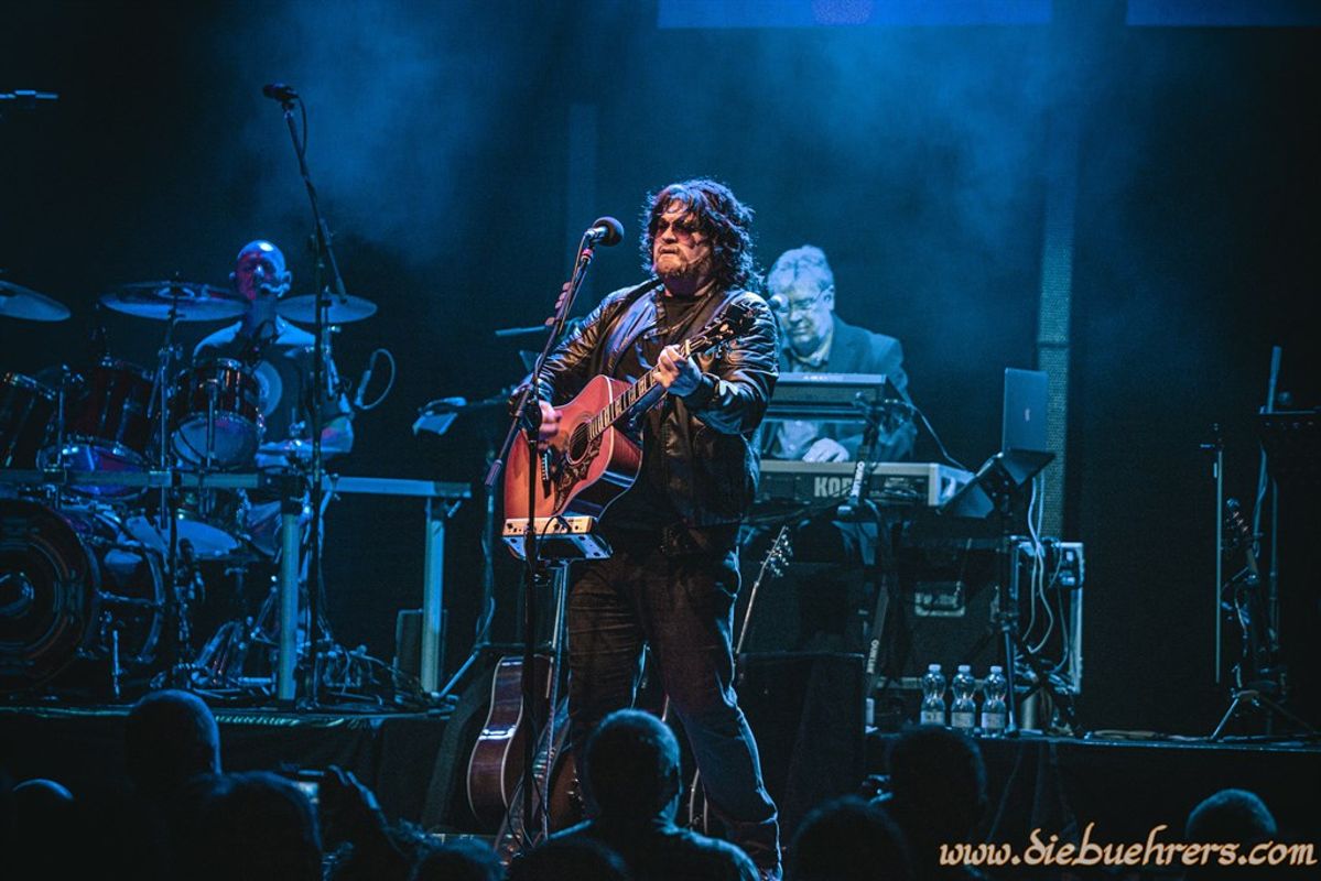 A musician in a leather jacket plays acoustic guitar under blue stage lights, flanked by a drummer and a keyboardist. The scene conveys energy and passion.