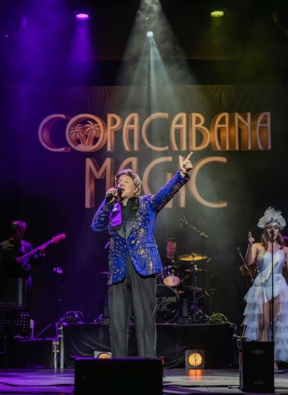 Martyn James in a glittery blue jacket performs energetically under a spotlight on stage, with a live band and backup singer. The backdrop reads "Copacabana Magic."