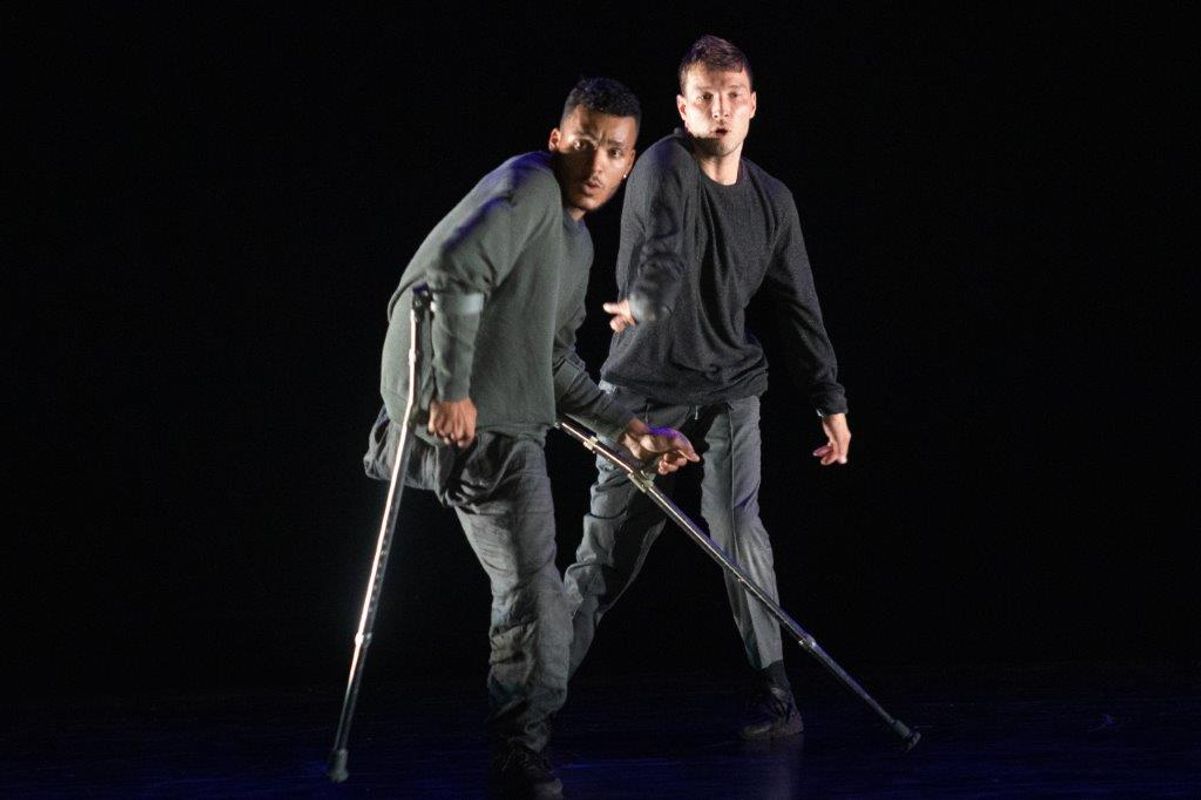 Two people in dark grey clothing dance on stage with one using crutches. 