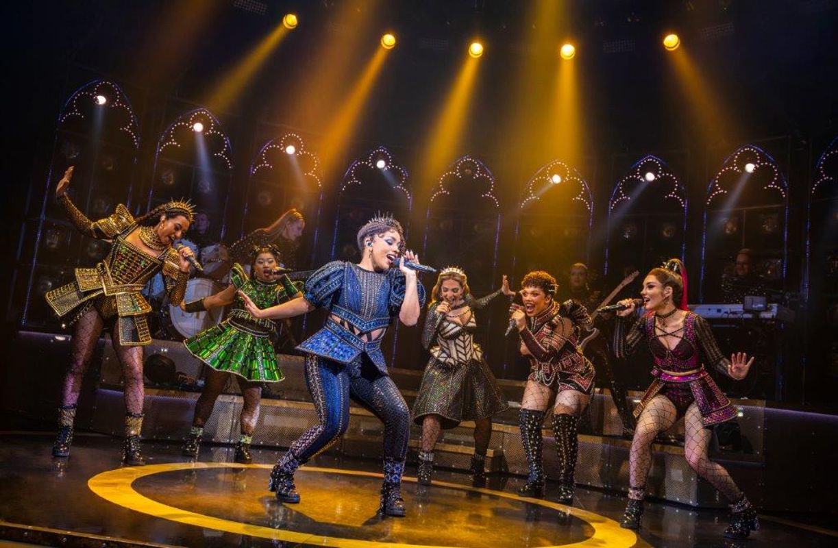 A person wearing a black and blue studded outfit stands centrally with five other people surrounding them.