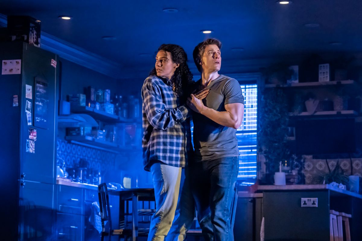Two people stand close together in a dimly lit kitchen, bathed in blue light, holding each other and looking anxiously off to the side as if reacting to something unseen. The space feels tense and eerie, with everyday kitchen items visible in the background.