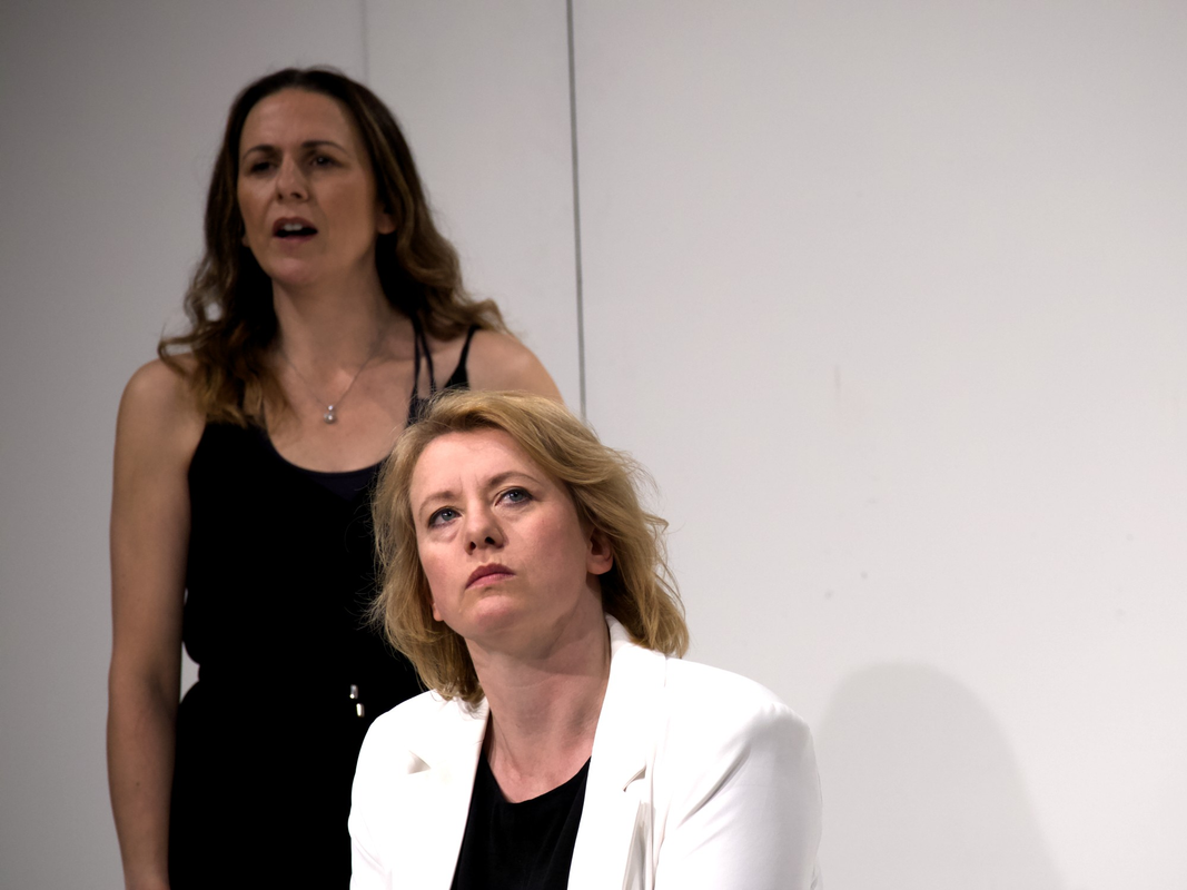 Two people in a white room, one is stood behind the other that is sitting in a chair