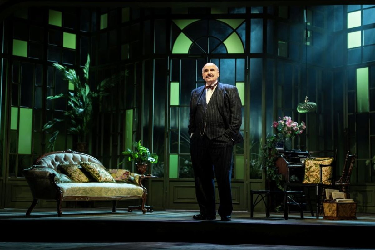 A person stands confidently on a well-lit theater stage set designed to evoke an elegant room, featuring a vintage sofa, plants, a small table with floral arrangements, and architectural elements with large windows.