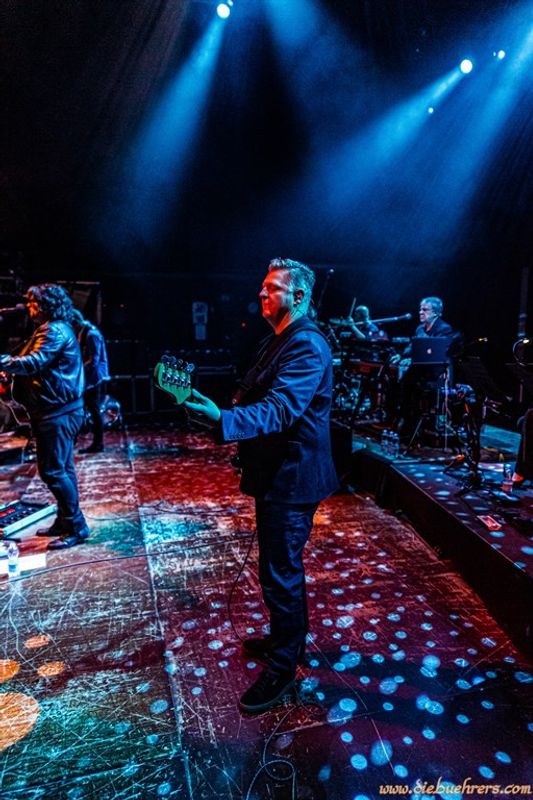 Live concert scene with musicians on stage. A guitarist stands in the foreground, while another musician plays keyboards under blue stage lights, creating an energetic atmosphere.